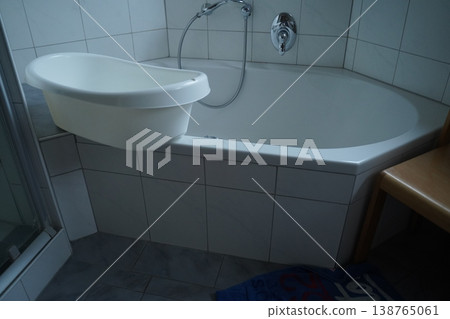 White bathtub with a curved wall-mounted sink and metal faucet in a tiled bathroom, with a shower panel on the left. Simple bathroom fixtures in a clean setting. 138765061