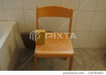 Wooden chair in a tiled bathroom with a yellow sponge on the seat beside a bathtub. Simple indoor scene shows cleaning item on chair Wooden chair in a tiled bathroom with a yellow sponge on the seat beside a bathtub. Simple indoor scene shows cleaning item on chair 138765065
