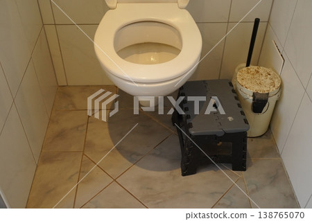 Toilet in a tiled bathroom with a plastic step stool and a covered bucket beside a cleaning brush. Simple restroom scene. Toilet in a tiled bathroom with a plastic step stool and a covered bucket beside a cleaning brush. Simple restroom scene. 138765070