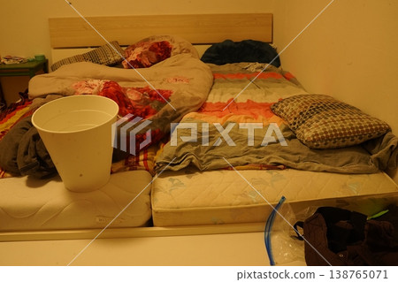 Messy bedroom with low bed and rumpled blankets, pillows, and stained fabric; a white bucket sits on a mattress with scattered clothes and bags. The room looks untidy. Messy bedroom with low bed and rumpled blankets, pillows, and stained fabric; a white bucket sits on a mattress with scattered clothes and bags. The room looks untidy. 138765071