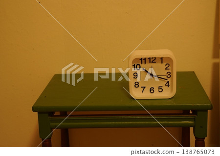 White analog alarm clock on a green wooden table against a beige wall, showing numbers "1 2 3 4 5 6 7 8 9 10 11 12". A simple indoor still life. 138765073