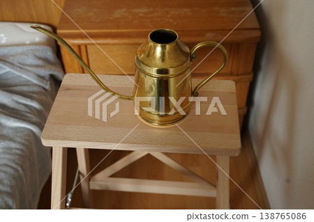 Brass watering can with long spout on a small wooden table beside a bed in a cozy room. Simple indoor still life with warm tones. Brass watering can with long spout on a small wooden table beside a bed in a cozy room. Simple indoor still life with warm tones. 138765086