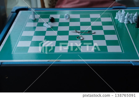 Green checkered game board with white pieces and a pair of scissors in the center; pieces clustered at the edge. A tabletop game scene. Green checkered game board with white pieces and a pair of scissors in the center; pieces clustered at the edge. A tabletop game scene. 138765091