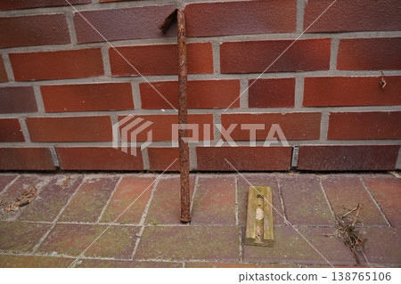 Rusty metal rod leaning against brick wall above tiled pavement with a small ground fixture and dry twigs 138765106