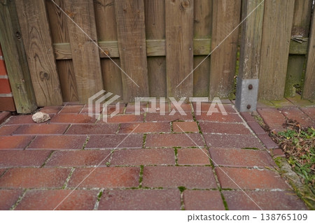 Brick patio paving with moss and weeds beside a wooden fence and metal post bracket Brick patio paving with moss and weeds beside a wooden fence and metal post bracket 138765109