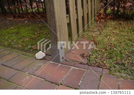 Wooden fence post base on brick paving with a white stone nearby; grass and fallen leaves beside the walkway Wooden fence post base on brick paving with a white stone nearby; grass and fallen leaves beside the walkway 138765111