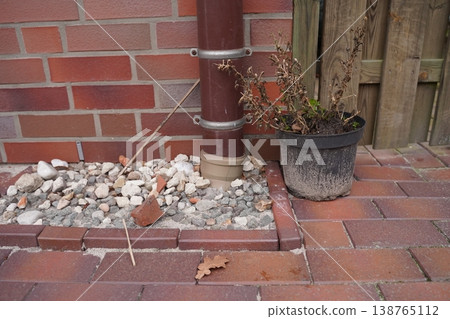 Brick wall with drainpipe, gravel bed, paving stones, and a flowerpot with dried plants beside a wooden fence 138765112