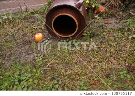 Brown ceramic jar lying on its side on patchy grass and soil, with a red apple nearby Brown ceramic jar lying on its side on patchy grass and soil, with a red apple nearby 138765117