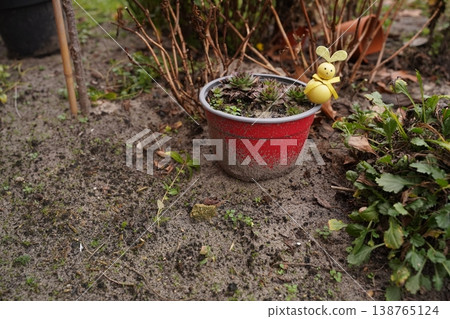 Red flowerpot with dried plant in sandy garden soil, small yellow insect decoration on rim, green foliage nearby Red flowerpot with dried plant in sandy garden soil, small yellow insect decoration on rim, green foliage nearby 138765124