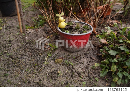Red flowerpot with dry plants and a small bunny figurine on sandy ground in a garden bed 138765125