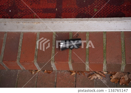 Small black plastic object lying on a brick step beside a metal threshold and a red doormat, with dry leaves nearby Small black plastic object lying on a brick step beside a metal threshold and a red doormat, with dry leaves nearby 138765147