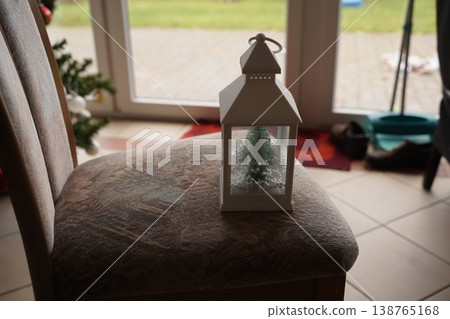 White lantern with small tree decoration on an upholstered chair near a glass door, soft indoor light, blurred patio view 138765168