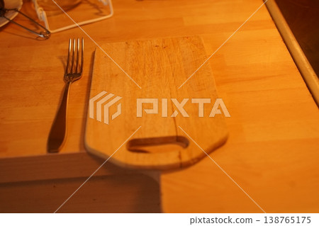 Wooden cutting board on a table with a fork beside it under warm light; blurred kitchen items in background 138765175