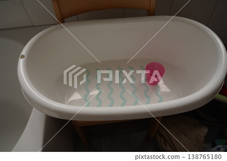 White baby bathtub on a stand with shallow water, blue wavy anti-slip lines, and a pink cup inside in a tiled bathroom 138765180