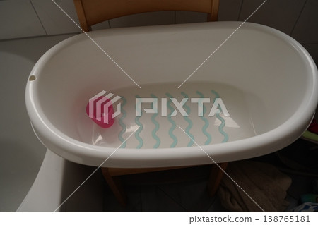White baby bathtub filled with water, a pink cup inside, and a mat with blue wavy lines on the bottom in a tiled bathroom 138765181