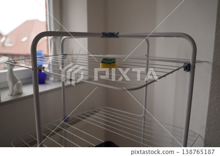 Metal drying rack near a window indoors, with a cleaning sponge on the top shelf and blurred buildings outside 138765187