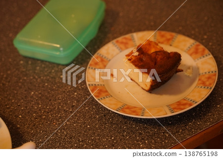 Slice of baked cake on a patterned plate on a kitchen countertop, with a closed plastic container nearby Slice of baked cake on a patterned plate on a kitchen countertop, with a closed plastic container nearby 138765198