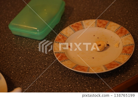 Plate with crumbs and a single ring-shaped snack on a speckled countertop beside a closed plastic container Plate with crumbs and a single ring-shaped snack on a speckled countertop beside a closed plastic container 138765199