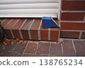 Blue wedge doorstop on brick doorstep by white sliding door, with fallen leaves on paved walkway 138765234