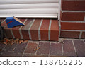 Small blue and red cardboard box on brick doorstep beside white siding and brick wall, with dry leaves on the ground 138765235