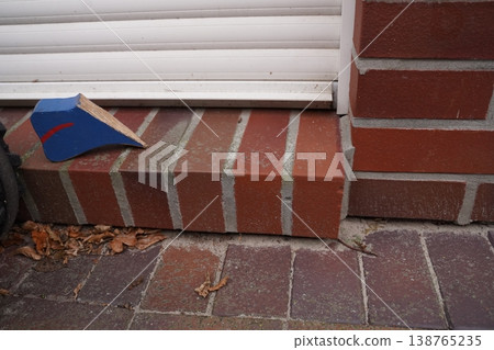 Small blue and red cardboard box on brick doorstep beside white siding and brick wall, with dry leaves on the ground 138765235
