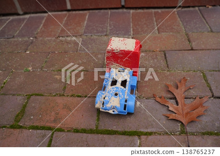 Small toy vehicle on brick pavement beside a dry leaf; red and blue plastic, worn and dirty, outdoors Small toy vehicle on brick pavement beside a dry leaf; red and blue plastic, worn and dirty, outdoors 138765237
