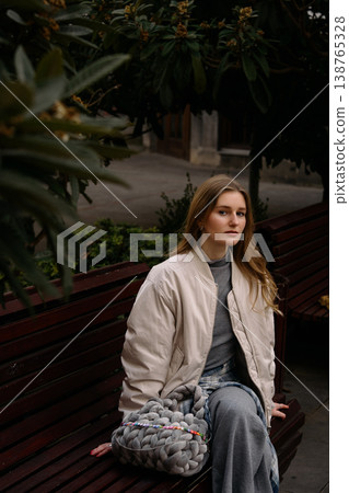 Young woman sits on a bench in a park wearing a casual outfit with a handbag Young woman sits on a bench in a park wearing a casual outfit with a handbag 138765328
