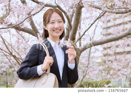 A young woman in a business suit, a new employee, strikes a triumphant pose under cherry blossoms – an image of spring job hunting. A young woman in a business suit, a new employee, strikes a triumphant pose under cherry blossoms – an image of spring job hunting. 138765687
