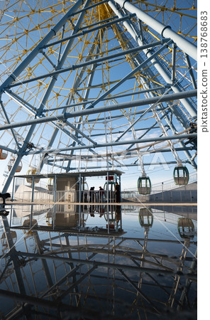 Ground level view of a Ferris wheel and its complex steel structure reflected in a large puddle 138768683