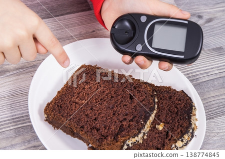 Hand of child holding glucometer, sweet chocolate cake. Checking sugar level during diabetes Hand of child holding glucometer, sweet chocolate cake. Checking sugar level during diabetes 138774845