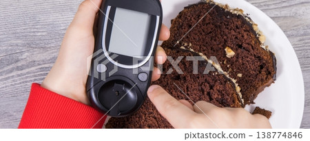 Hand of child holding glucometer, sweet chocolate cake. Checking sugar level during diabetes Hand of child holding glucometer, sweet chocolate cake. Checking sugar level during diabetes 138774846