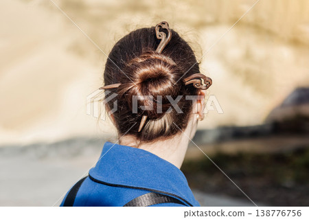 Woman with neat hair bun secured by wooden hairpin wearing a blue coat photographed from behind 138776756