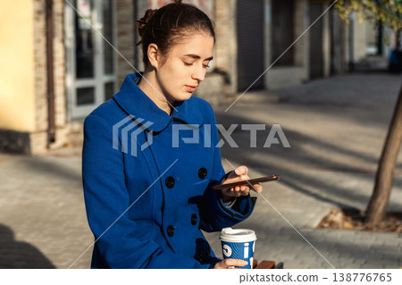 Young woman in blue coat checking phone while holding coffee on a city bench Young woman in blue coat checking phone while holding coffee on a city bench 138776765