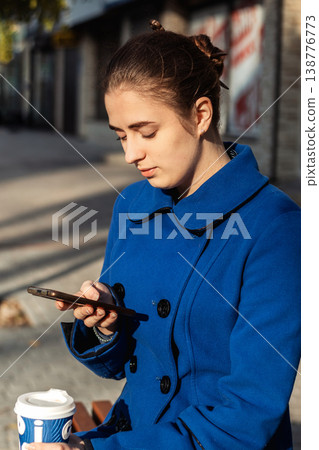 Young woman in a blue coat checking her smartphone while holding a takeaway coffee on a city bench Young woman in a blue coat checking her smartphone while holding a takeaway coffee on a city bench 138776773