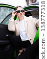 Stylish woman in sunglasses stepping out of a green car in an elegant beige coat 138777318