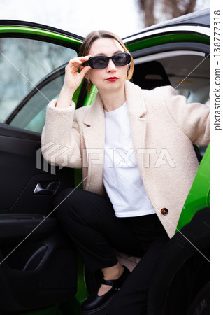 Stylish woman in sunglasses stepping out of a green car in an elegant beige coat Stylish woman in sunglasses stepping out of a green car in an elegant beige coat 138777318