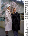 Two caucasian women dressed in stylish coats are smiling and walking outdoors in an urban area. 138777352