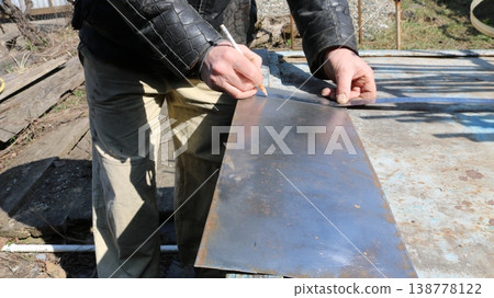 marking and checking the required size on a small sheet of metal lying on the work surface in an outdoor craft workshop using a pencil and ruler in the hands of a craftsman, marking an iron plate marking and checking the required size on a small sheet of metal lying on the work surface in an outdoor craft workshop using a pencil and ruler in the hands of a craftsman, marking an iron plate 138778122
