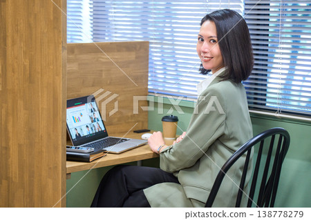 一位女商人坐在靠窗的隔間座位上，側身觀看線上會議。 138778279