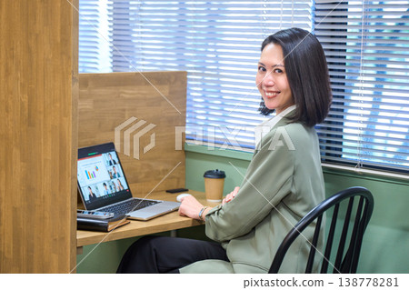 一位女商人坐在靠窗的隔間座位上，側身觀看線上會議。 138778281