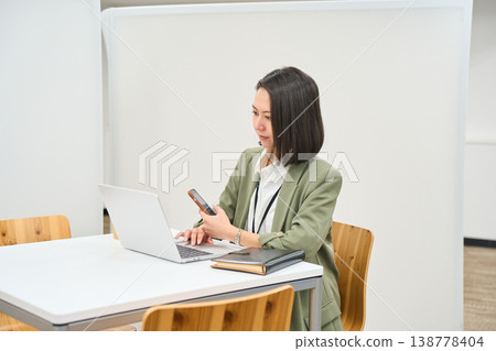 一名女員工在辦公桌前打開筆記型電腦,並操作她的智慧型手機。 一名女員工在辦公桌前打開筆記型電腦,並操作她的智慧型手機。 138778404