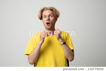 Young man standing against a neutral background, showing a strong reaction of fright and disbelief with his eyes wide open, mouth agape, and hands clenched up near his neck 138779849