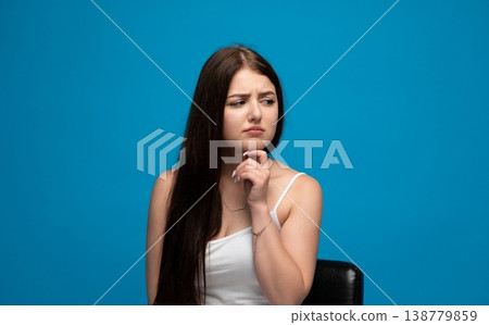 Young woman thinking, looking doubtful, holding chin, contemplating decisions on blue background Young woman thinking, looking doubtful, holding chin, contemplating decisions on blue background 138779859