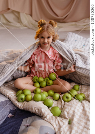 Little girl sitting on mattress with green apples, wrapped in blanket, studio drapery background Little girl sitting on mattress with green apples, wrapped in blanket, studio drapery background 138780366