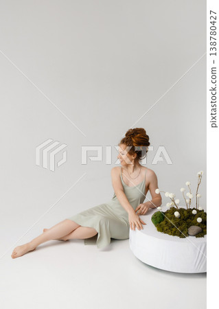 Young Caucasian woman in light dress touching dried flowers on moss on white pedestal, white wall Young Caucasian woman in light dress touching dried flowers on moss on white pedestal, white wall 138780427