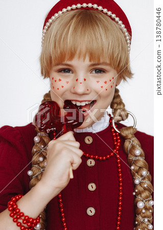 Smiling Caucasian girl eating rooster lollipop in red folk dress on light background Smiling Caucasian girl eating rooster lollipop in red folk dress on light background 138780446