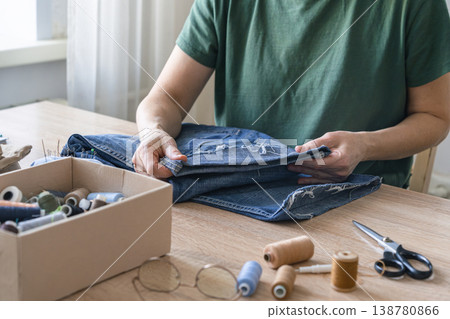 Person hands holding and mending ripped denim jeans on a wooden table, embracing eco-friendly repair and upcycling, for sustainable fashion. Reusing things 138780866