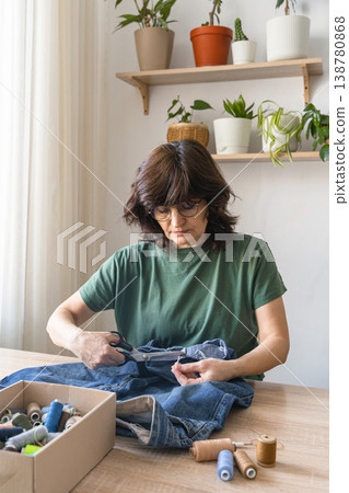 An adult woman cutting thread from old jeans for reuse, promoting sustainable fashion and circular economy principles. Person upcycling denim jeans with scissors, repairing clothing 138780868