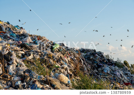 A large garbage landfill site shows piles of waste under a clear blue sky with birds flying overhead in the distance A large garbage landfill site shows piles of waste under a clear blue sky with birds flying overhead in the distance 138780939