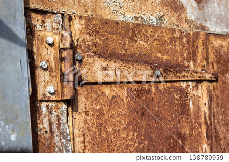Rusty metal door of a garage shows weathering and wear. The hinge and surface are visible against the background of a clear sky Rusty metal door of a garage shows weathering and wear. The hinge and surface are visible against the background of a clear sky 138780959